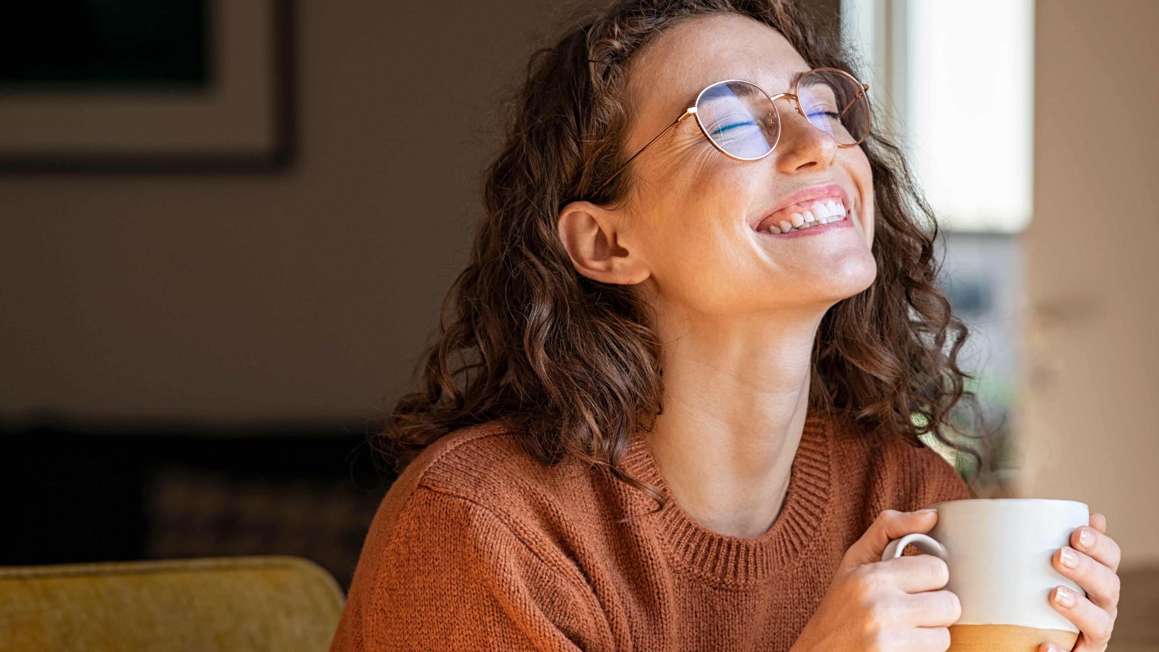 Woman wearing glasses and a brown sweater holding a white mug, smiling.