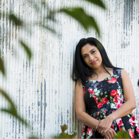 Woman - Reshmi Kumar in a floral dress standing against a textured wall with greenery.